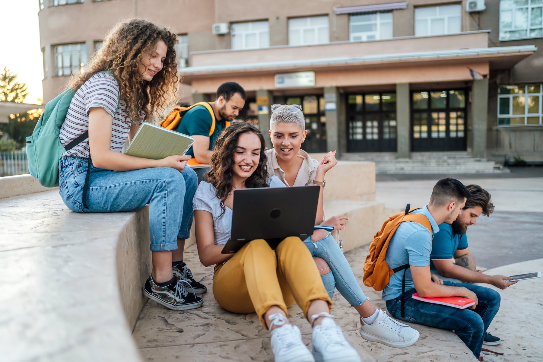 A group of female students sat on some steps outside talking and looking at a laptop A group of female students sat on some steps outside talking and looking at a laptop